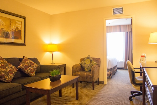 A hotel room with a sofa, armchair, dining table, and a small desk with a lamp.