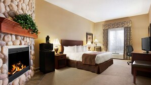 A hotel room with a stone fireplace, a bed with a brown bedspread, two bedside tables with lamps, a television, and a large window with curtains. - Days Inn & Suites by Wyndham Strathmore (Strathmore)