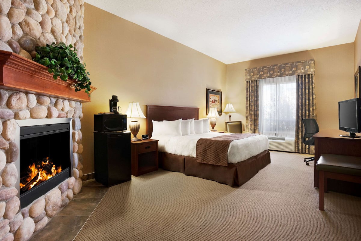 Une chambre d’hôtel avec une cheminée en pierre, un lit recouvert d’une courtepointe marron, deux tables de chevet avec des lampes, une télévision et une grande fenêtre avec des rideaux.