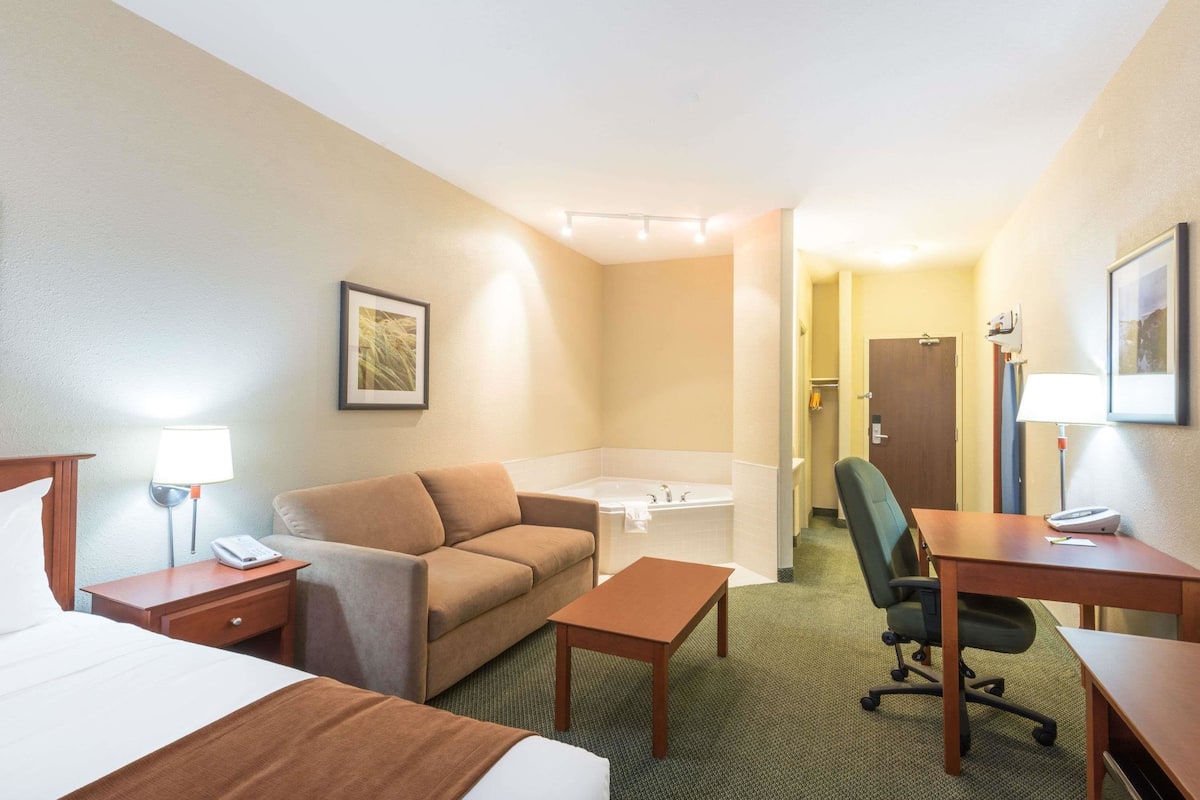 Habitación de hotel con cama, sofá, escritorio, silla, mesa, lámpara, teléfono y baño con bañera e inodoro.