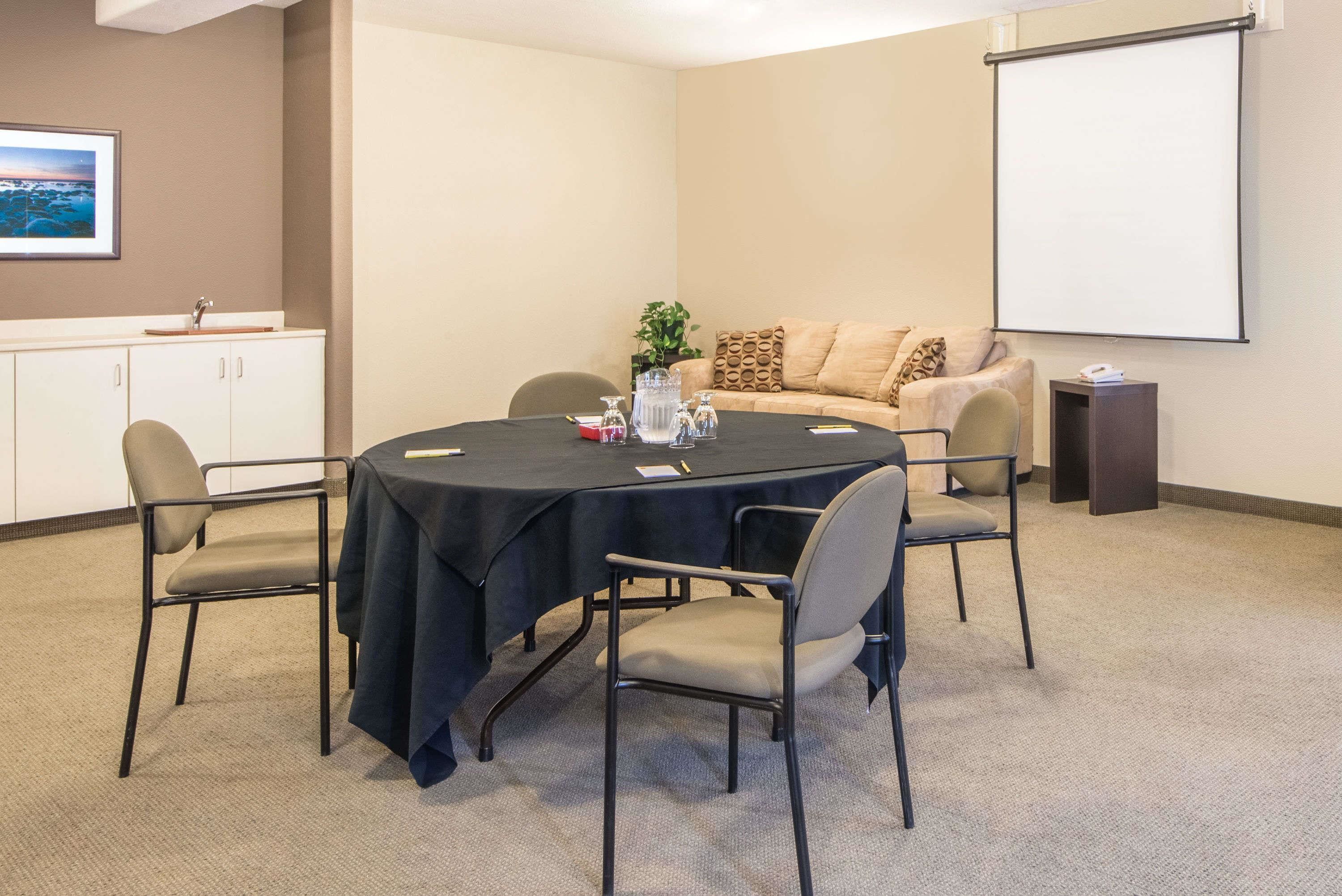Une salle de conférence équipée d’une table ronde, de quatre chaises, d’un écran de projection, d’un canapé et d’un meuble avec un évier.
