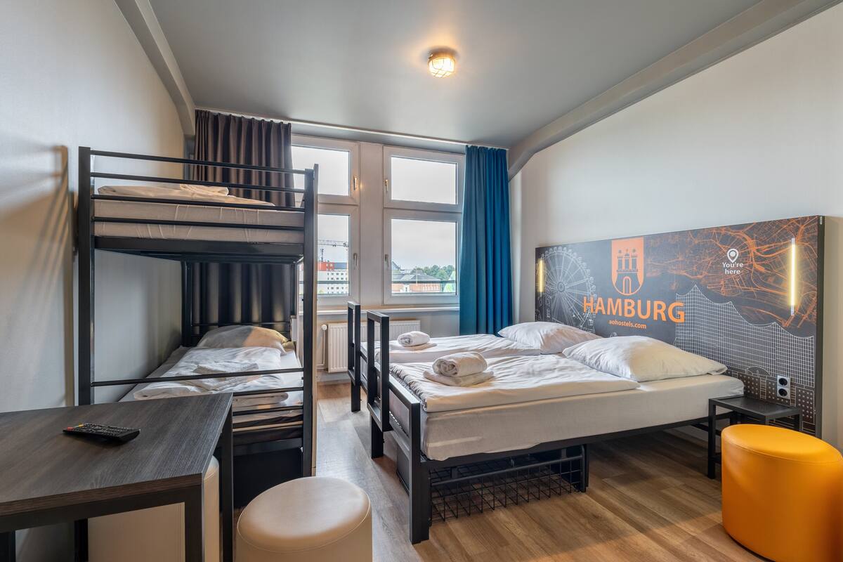 Ein Hotelzimmer mit Etagenbett, einem Einzelbett, einem Schreibtisch und einem Stuhl. Es gibt ein Fenster mit Vorhängen, eine Wand mit einem Stadtlandschaftsposter und einen kleinen, orangefarbenen Pouf.