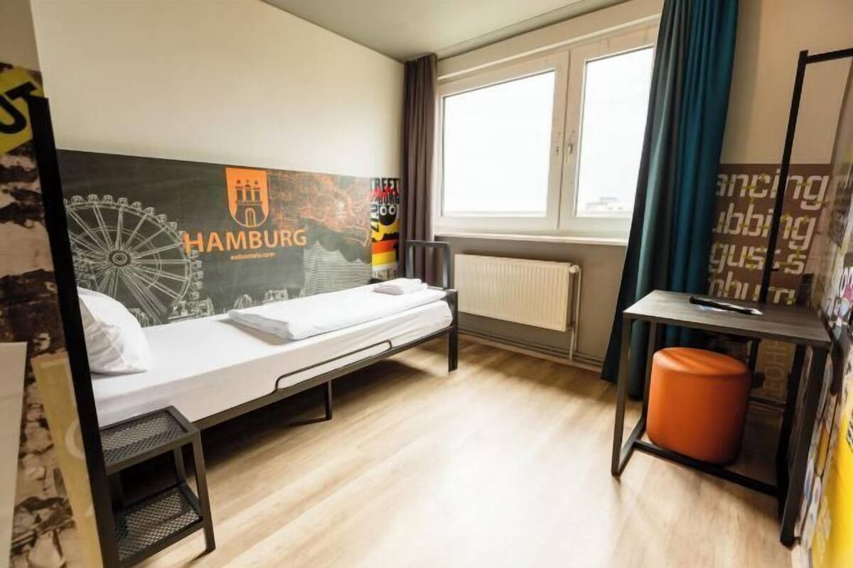 Ein Einzelzimmer mit Bett, Schreibtisch, Stuhl, Heizkörper, einem Fenster mit Vorhängen und einer Wand mit Stadtansicht und Text.