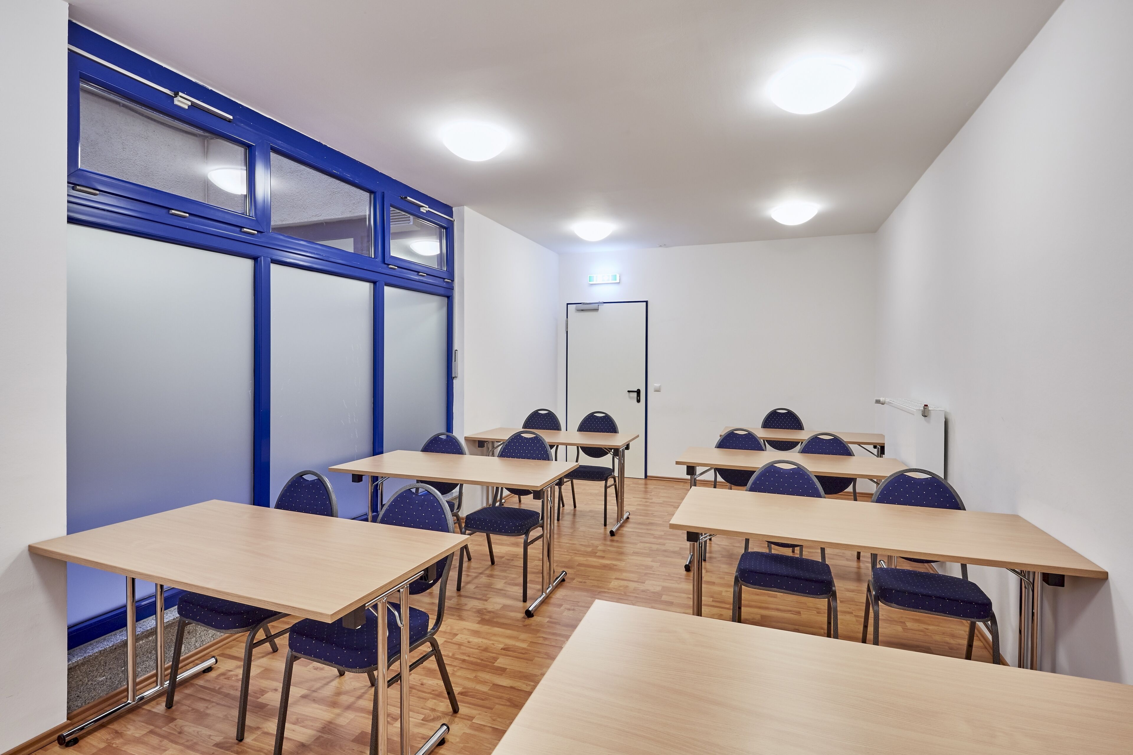 A classroom with wooden desks and chairs, white walls, and blue-framed windows.