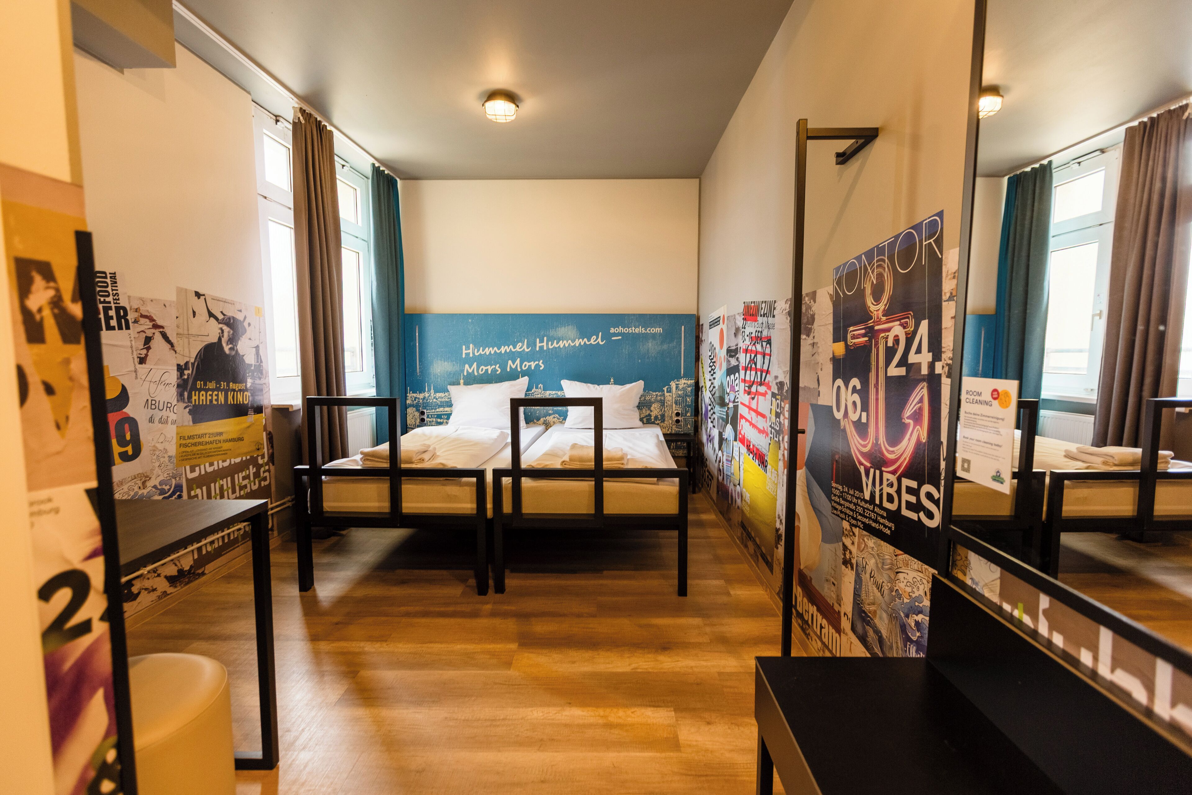 A hotel room with two beds, wooden flooring, a large mirror, and posters on the walls.