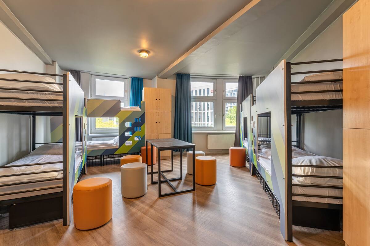 Ein Schlafsaalzimmer mit Etagenbetten, einem zentralen Tisch und orangefarbenen Hocker.