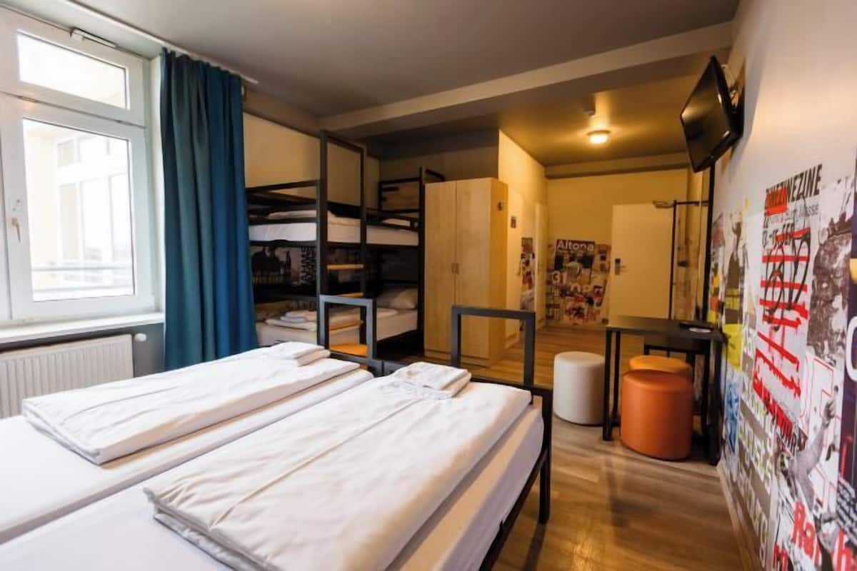 Ein Zimmer mit Etagenbetten, einem Vorhangfenster, einem kleinen Tisch und einer Wand mit Postern.
