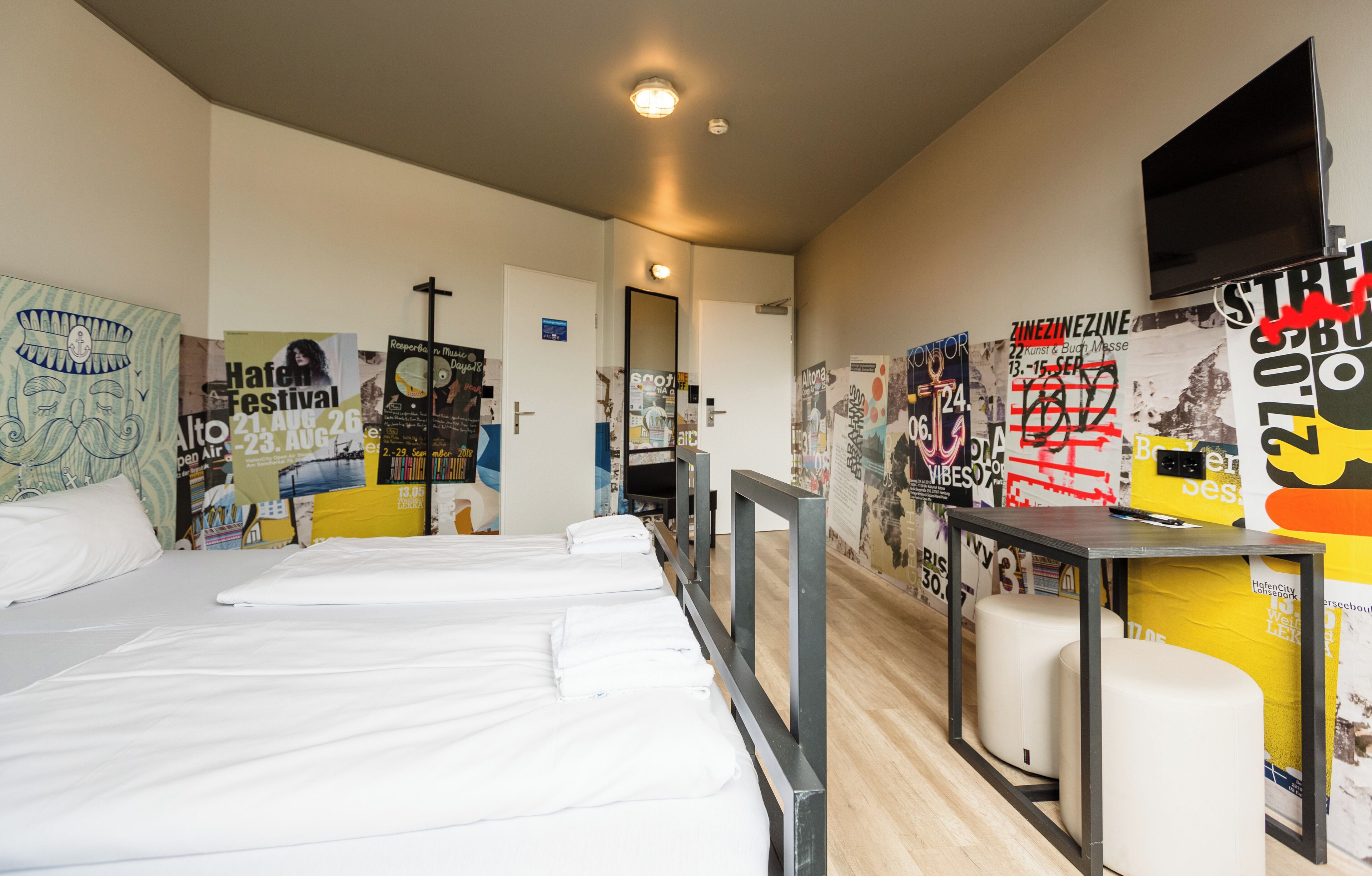 Habitación de hotel con dos camas, una mesita, un televisor y paredes decoradas con carteles y obras de arte.