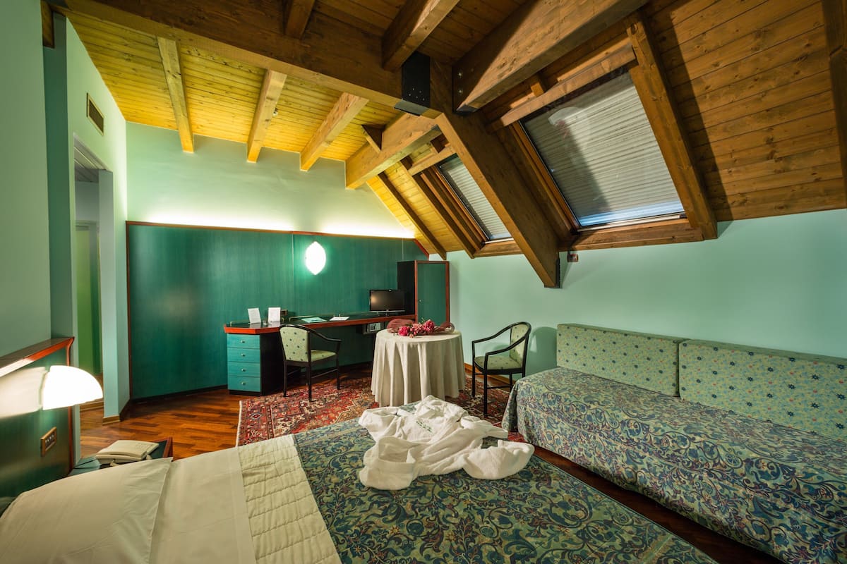 Ein Zimmer mit Bett, Schreibtisch, Stuhl, kleinem Tisch und Fenster mit Holzbalken.