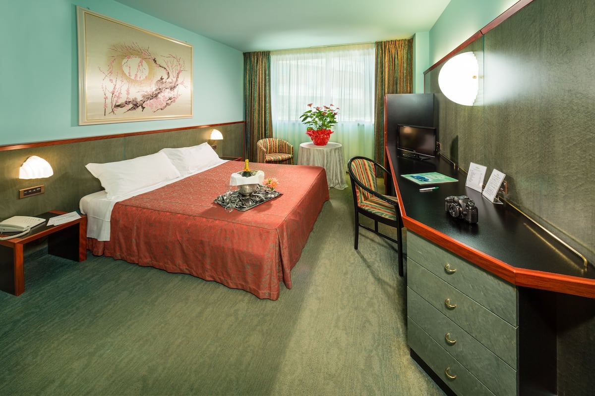 Ein Hotelzimmer mit Bett, Schreibtisch, Stuhl, Fernseher und einem Gemälde an der Wand.