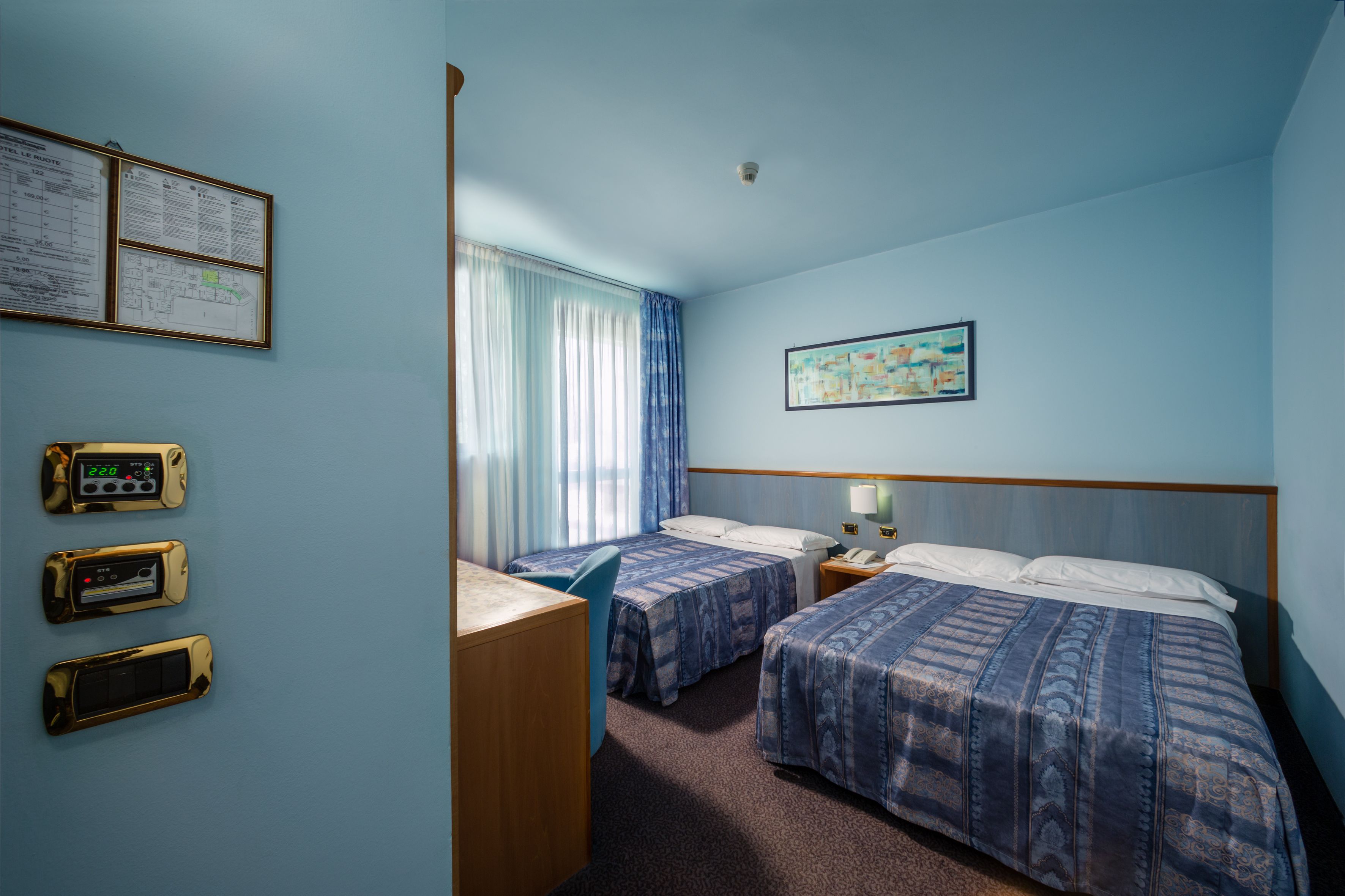 A hotel room with two beds, a desk, a chair, a nightstand, a lamp, a clock, and a map on the wall.