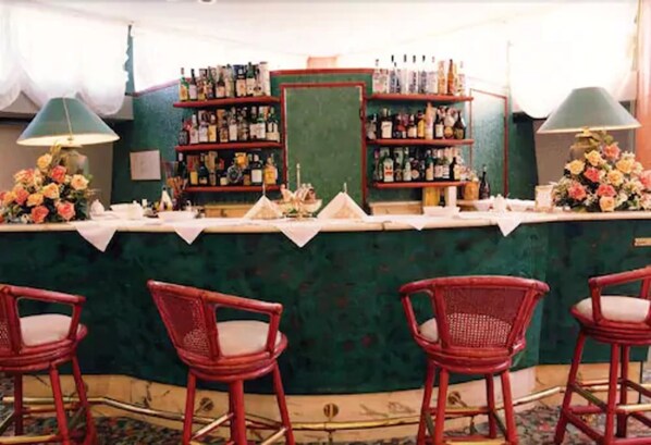 Zona de bar con sillas de mimbre, una barra verde y estantes llenos de botellas.