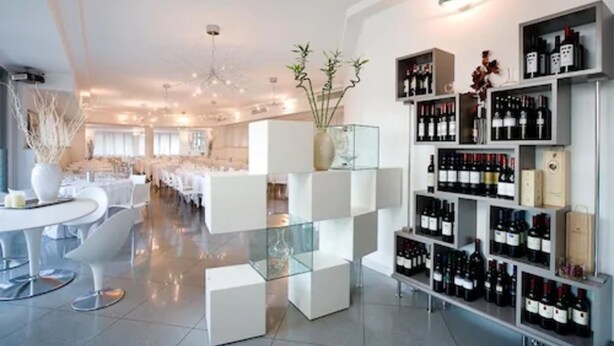 Un hall d’hôtel moderne avec une vitrine exposant des bouteilles de vin, des meubles blancs et un vase décoratif central.