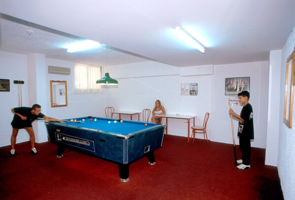 A pool table in a room with red carpet, white walls, and a ceiling fan. There are two people playing pool, and a third person sitting at a table.
