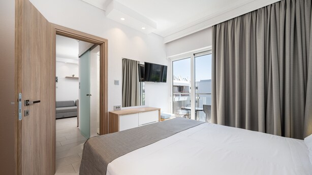 A modern hotel room with a large bed, a desk, a flat-screen TV, and a balcony with curtains. - Poseidon Hotel and Apartments (Kos)