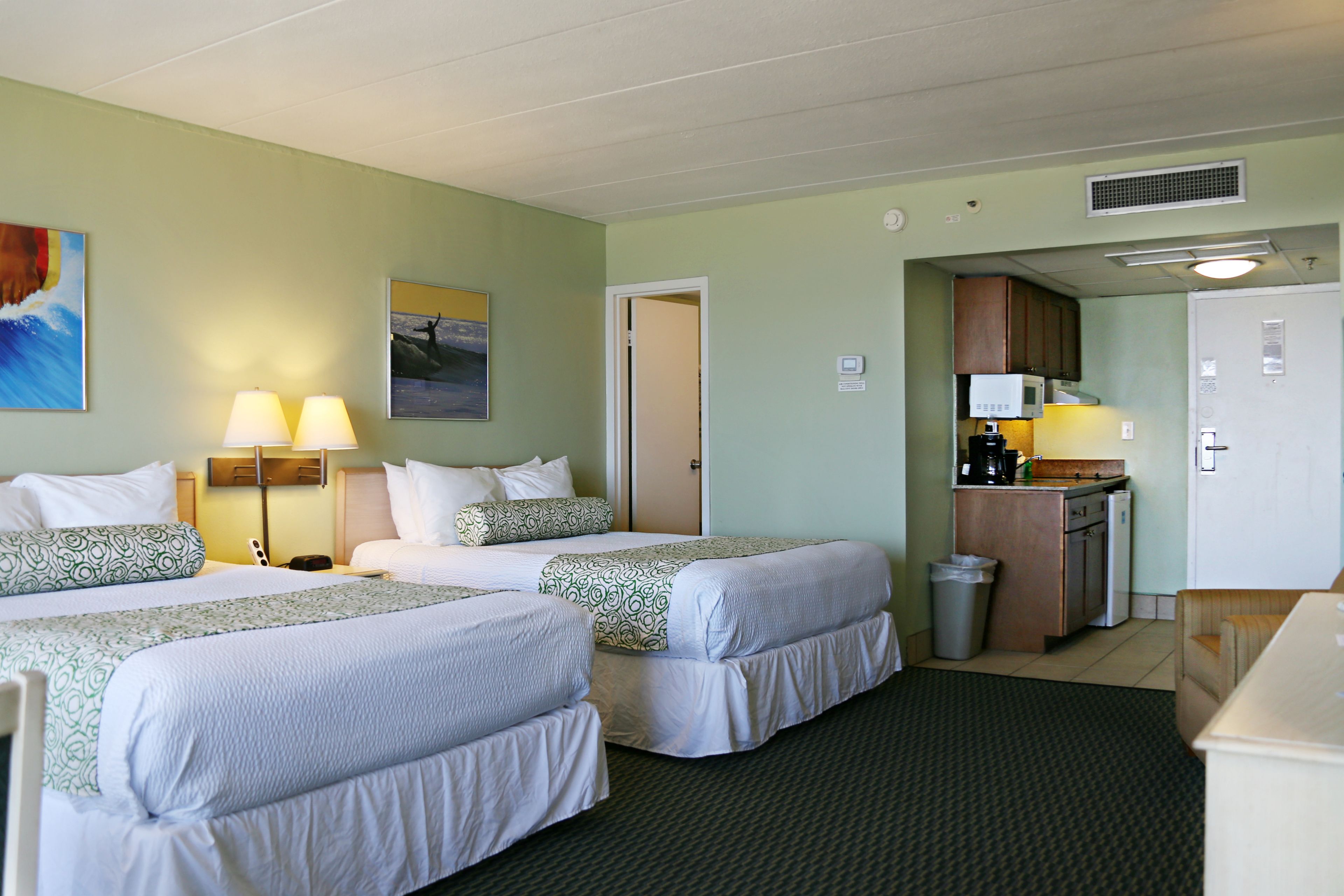 A hotel room with two beds, a kitchenette, a trash can, and a door leading to another room.