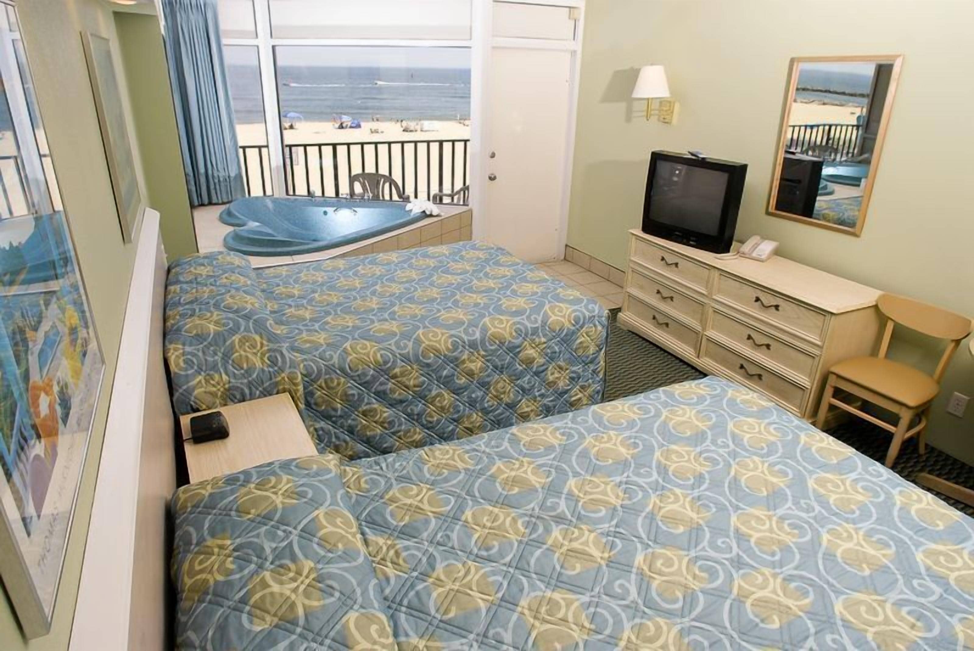 A hotel room with two beds, a TV, a desk, and a view of the beach.