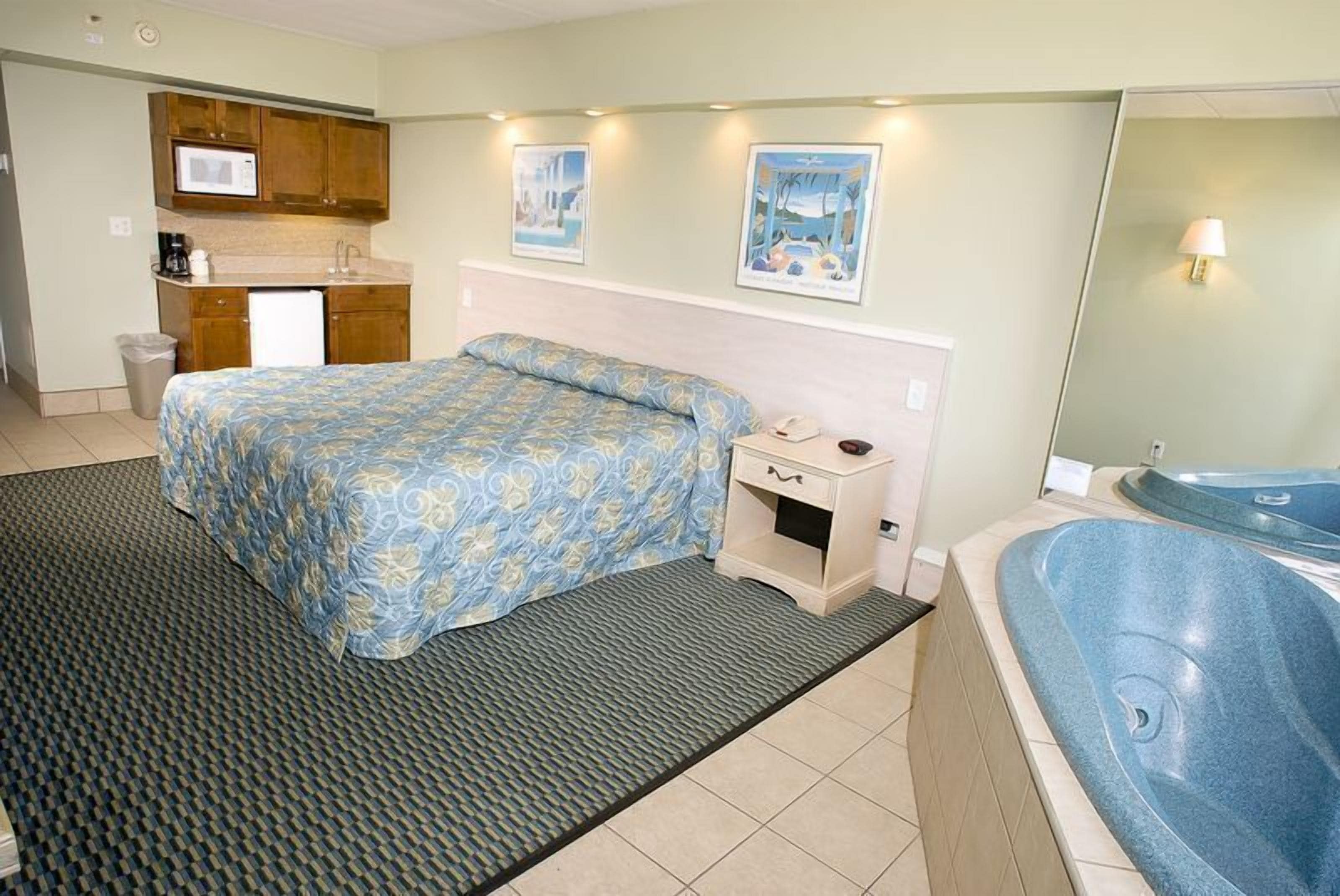 A hotel room with a bed, a nightstand, a small kitchenette, a bathroom with a bathtub, and artwork on the walls.