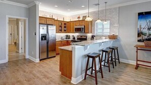 A modern kitchen with wooden cabinets, a stainless steel refrigerator, a large island with stools, and pendant lighting. - Beachgate CondoSuites and Oceanfront Resort (Port Aransas)