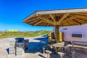 A covered outdoor seating area with a wooden table and benches, a picnic table, and a wooden structure with a roof.