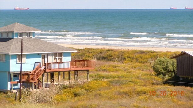 A coastal house with a balcony overlooking the ocean, a sandy beach in the foreground, and two ships in the distance. - Beachgate CondoSuites and Oceanfront Resort (Port Aransas)