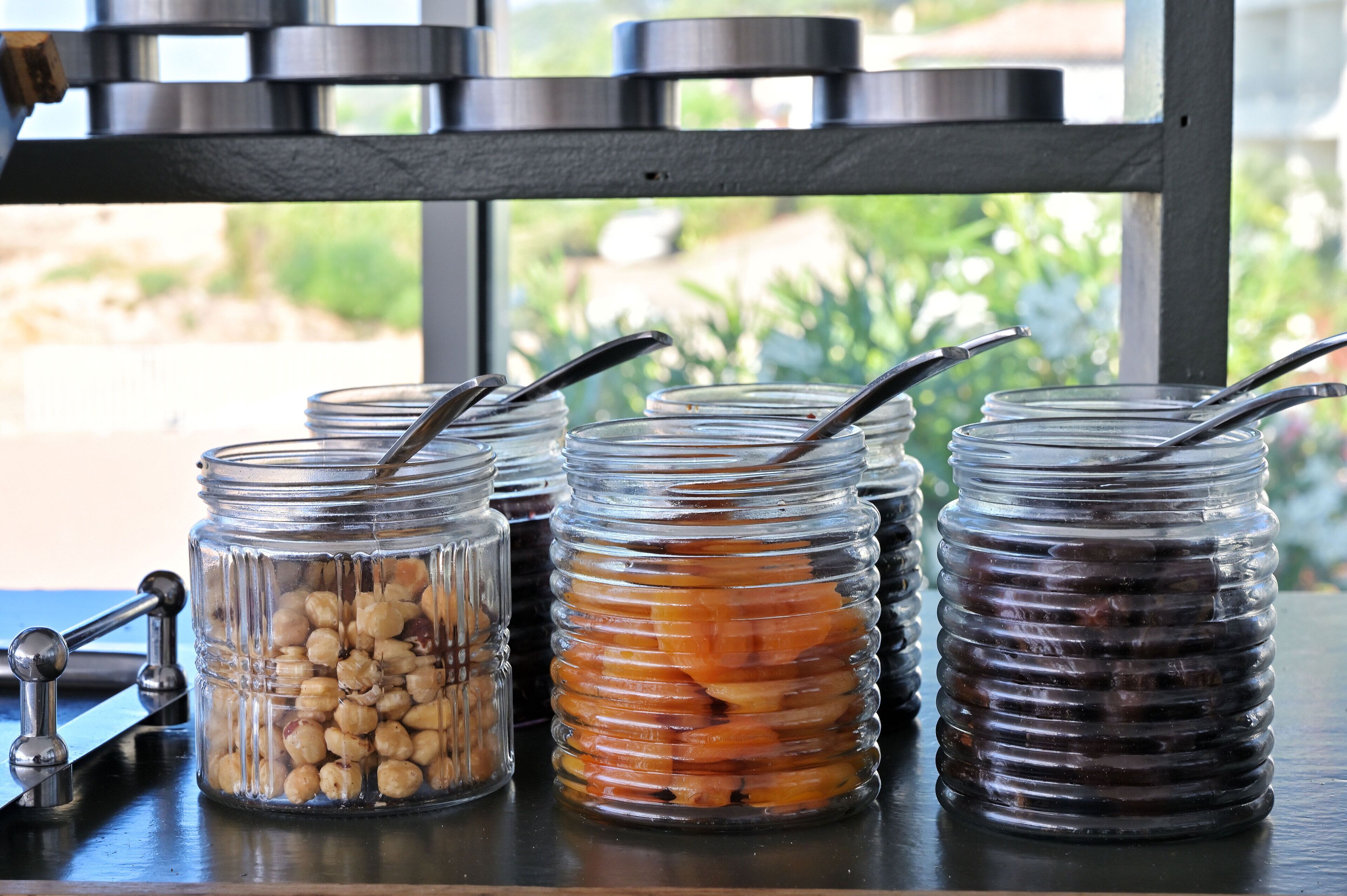Four glass jars with metal lids and spoons, containing different food items, placed on a table with a metal rack in the background.