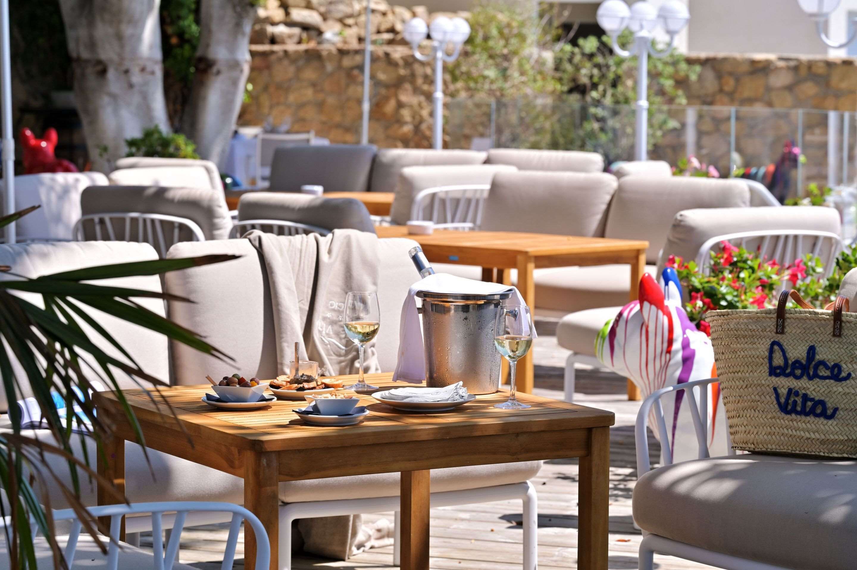 An outdoor seating area with wooden tables and white chairs, a metal bucket, and a wicker bag labeled "Dolce Vita".