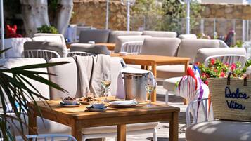 An outdoor seating area with wooden tables and white chairs, a metal bucket, and a wicker bag labeled "Dolce Vita".
