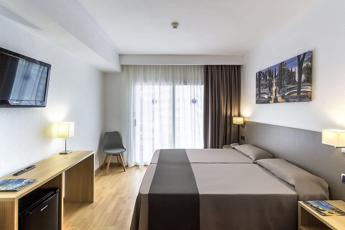 A hotel room with a bed, a desk with a TV, a chair, and a painting on the wall.