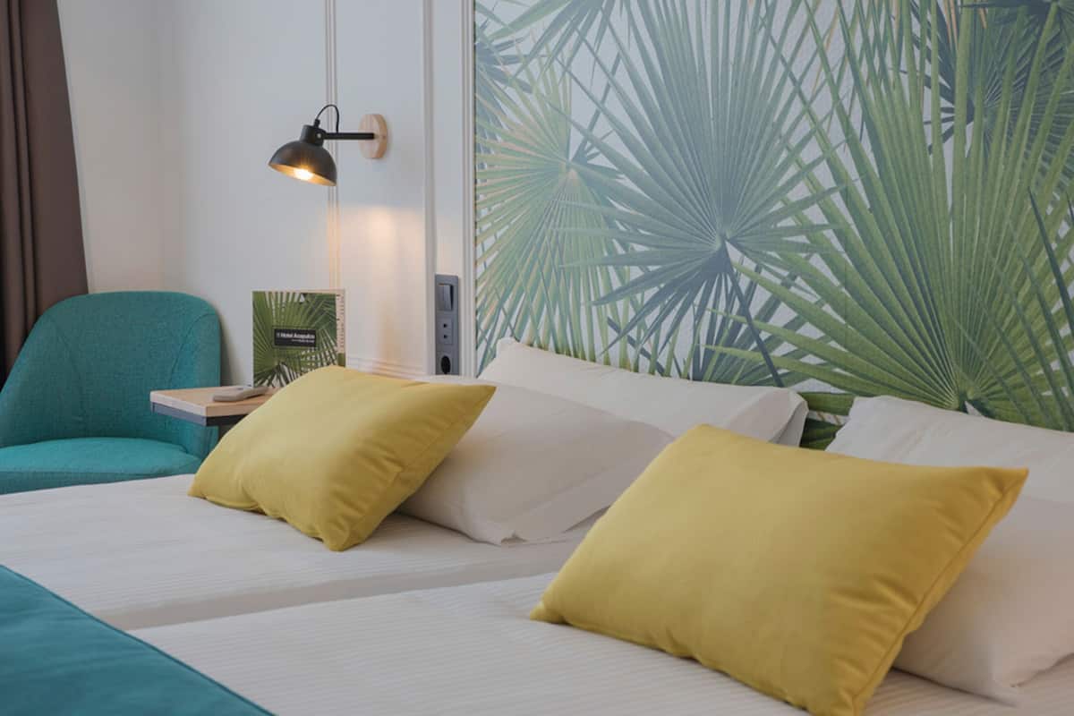 A hotel room with a bed, two pillows, a nightstand, a lamp, and a wall with a palm leaf pattern.