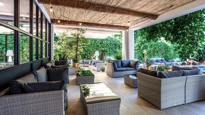A covered outdoor seating area with wicker furniture, large windows, and a view of a pool and greenery.