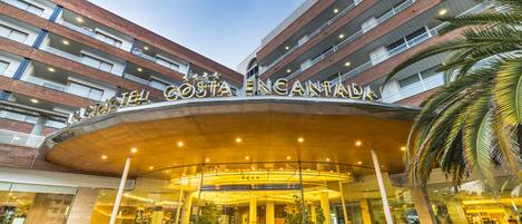 Entrada de un hotel con fachada curva, varios pisos y una palmera a la derecha.