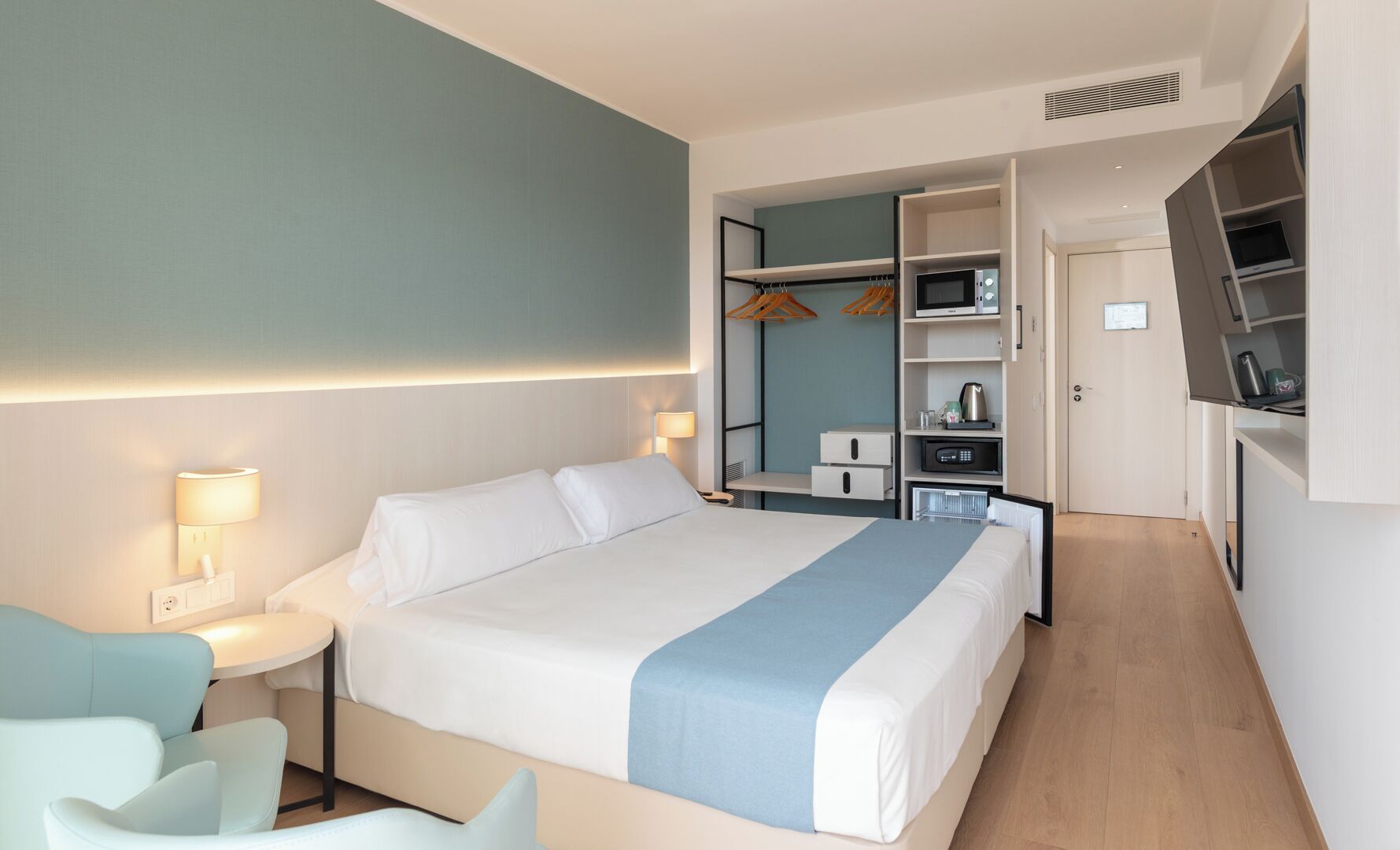 Habitación de hotel con cama, mesita de noche, lámpara, zona de estar, armario y kitchenette.