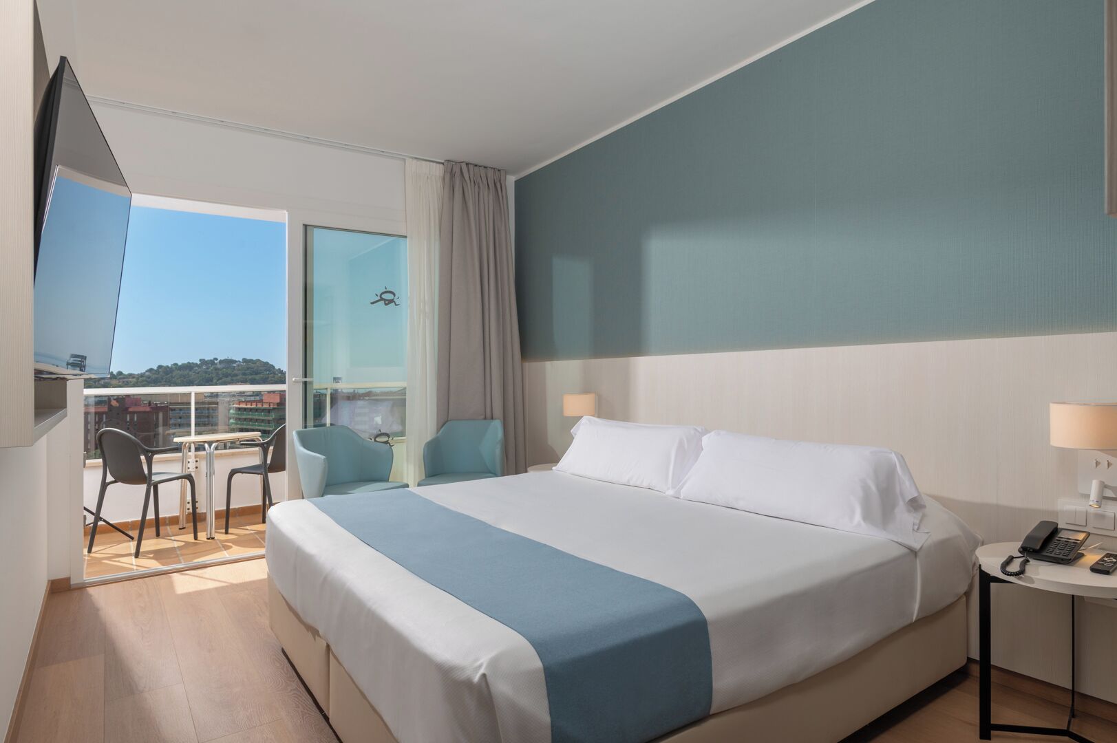 Habitación de hotel con cama, escritorio con teléfono, silla, balcón con mesa y sillas, y vista a edificios.
