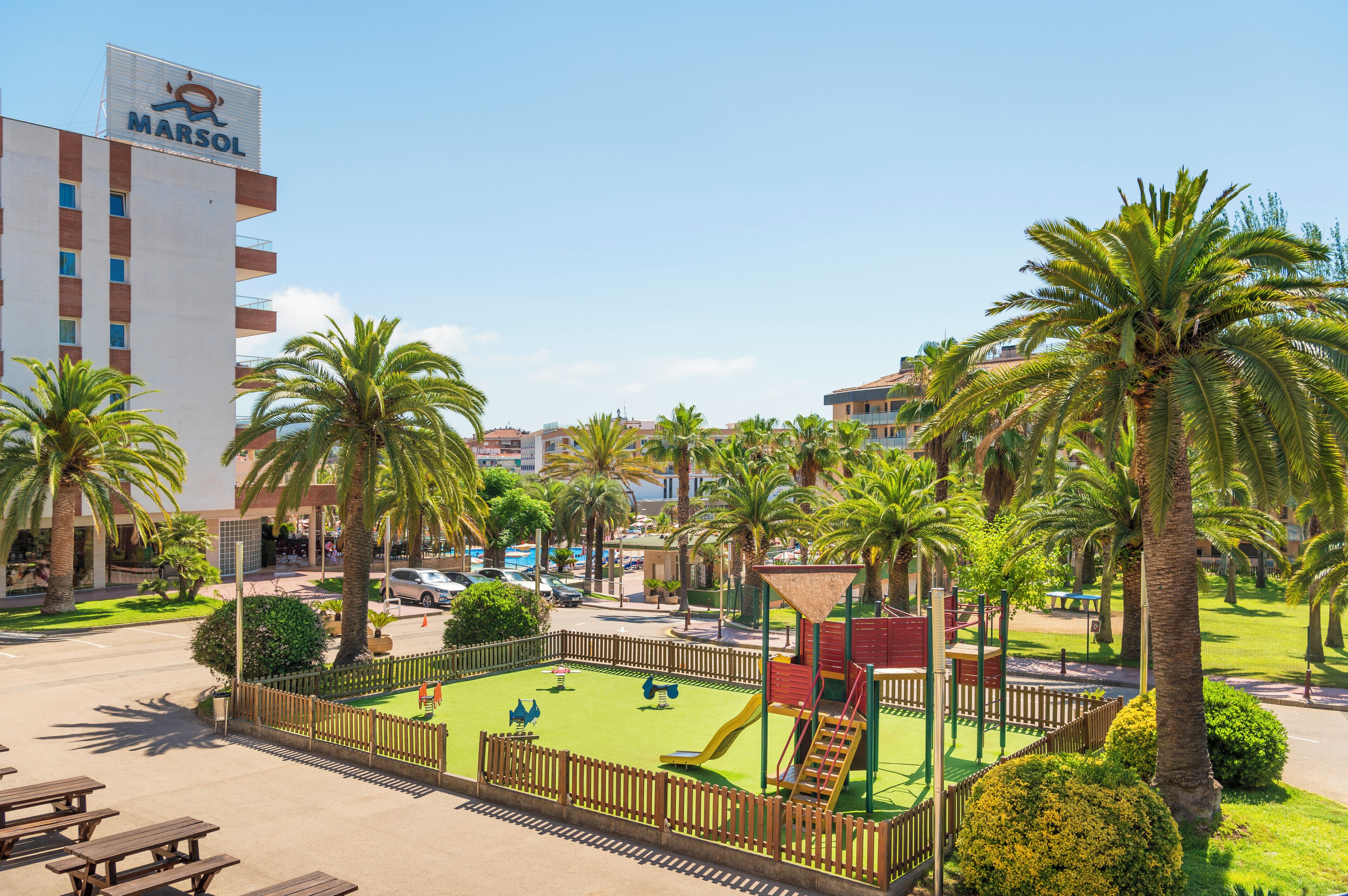 A hotel with a playground, palm trees, and a parking area.