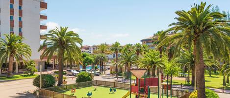 A hotel with a playground, palm trees, and a parking area.