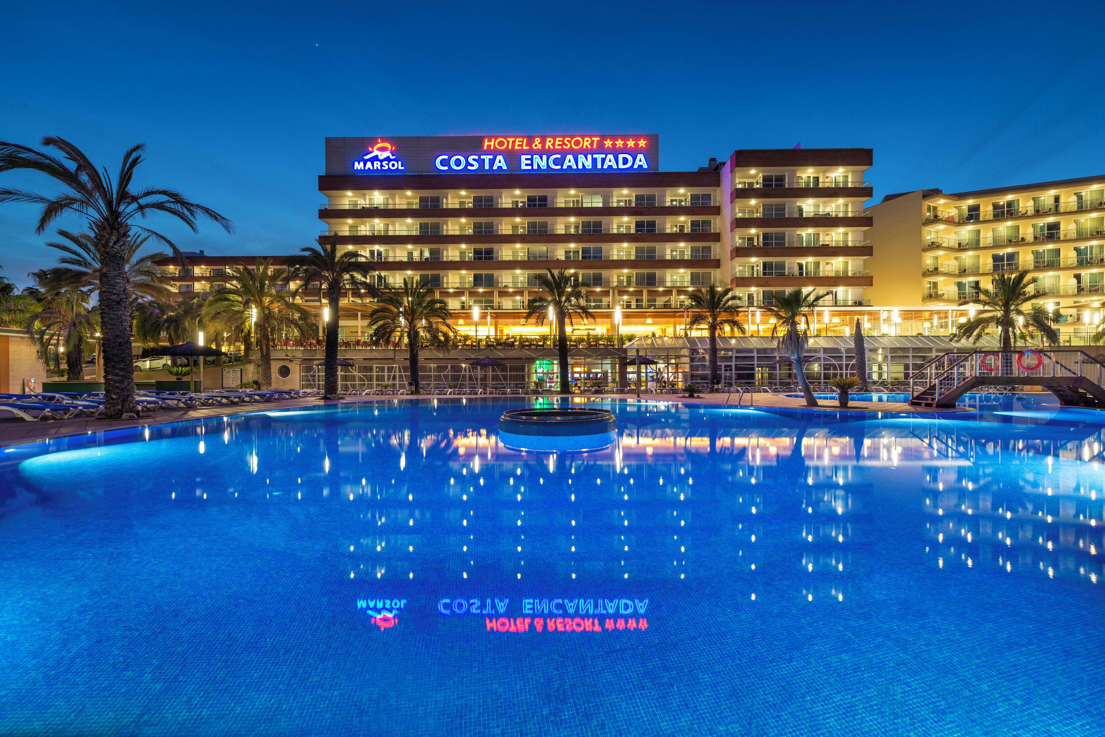 Een hotelzwembad met een bord waarop staat 'MARSOL COSTA ENCANTADA' en een groot gebouw met meerdere balkons op de achtergrond.