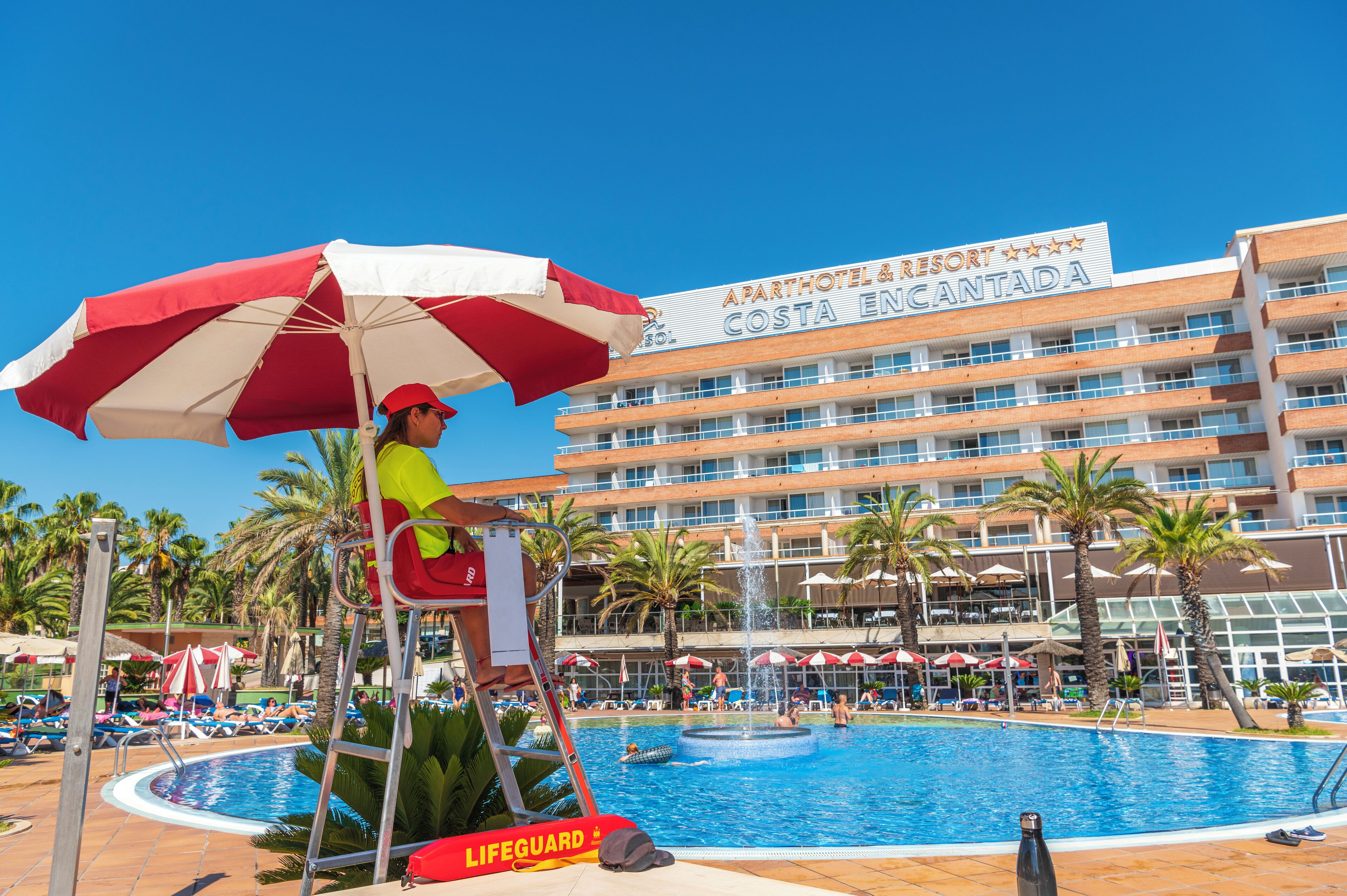 Ein Rettungsschwimmerstuhl mit einem rot-weißen Sonnenschirm, ein Schwimmbecken und ein Gebäude mit der Aufschrift 'APARTHOTEL & RESORT COSTA ENCANTADA'.