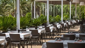 An outdoor dining area with multiple tables and chairs, surrounded by lush greenery and palm trees. - Costa Encantada Suites & Resort (Lloret de Mar)
