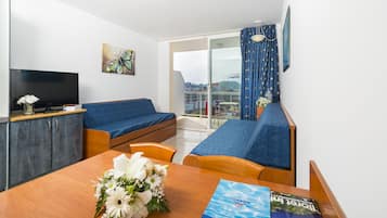 A modern hotel room with a balcony, a television, a sofa, a dining table with flowers, and a magazine.