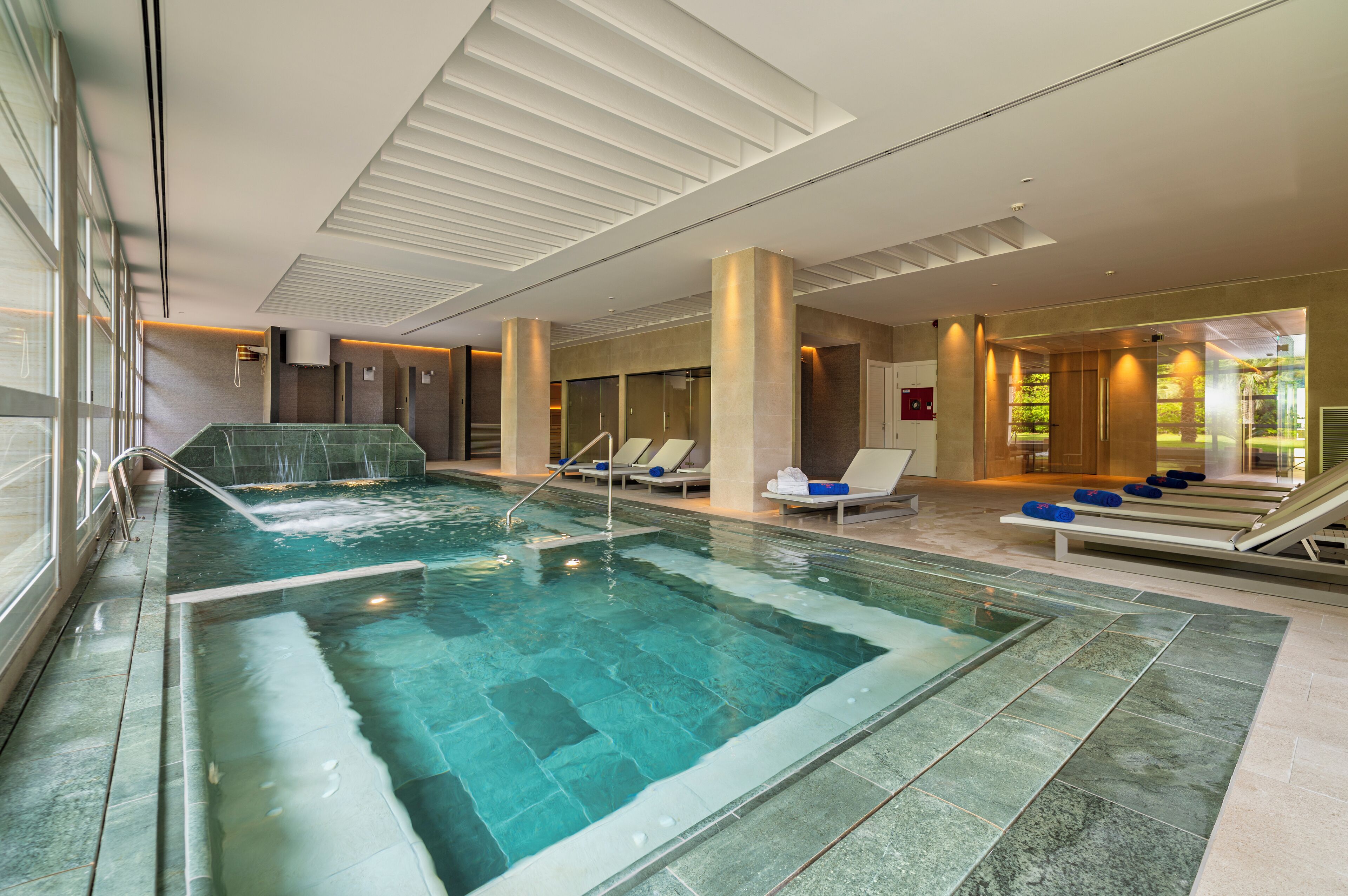 A modern indoor swimming pool with a waterfall feature, lounge chairs, and a view of the outdoors.