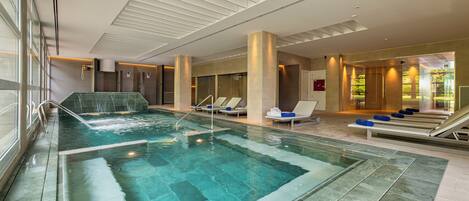 A modern indoor swimming pool with a waterfall feature, lounge chairs, and a view of the outdoors.