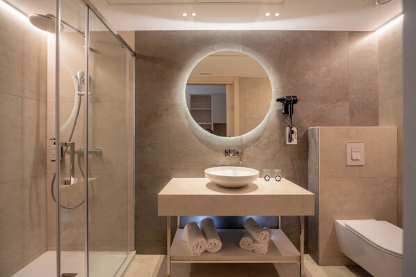 A modern bathroom with a glass-enclosed shower, a round mirror, a sink with a bowl, and a toilet. - Costa Encantada Suites & Resort (Lloret de Mar)