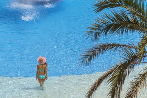 A person in a bikini walking in a clear blue pool with a palm tree in the foreground. - Costa Encantada Suites & Resort (Lloret de Mar)