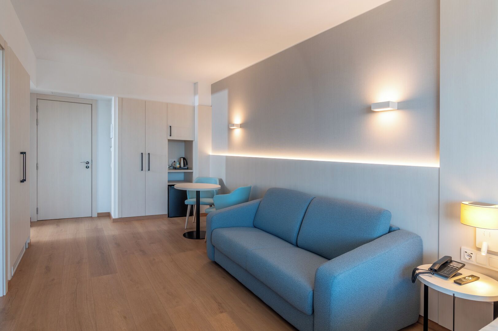 A modern hotel room with a sofa, a small table, a lamp, and a kitchenette in the background.