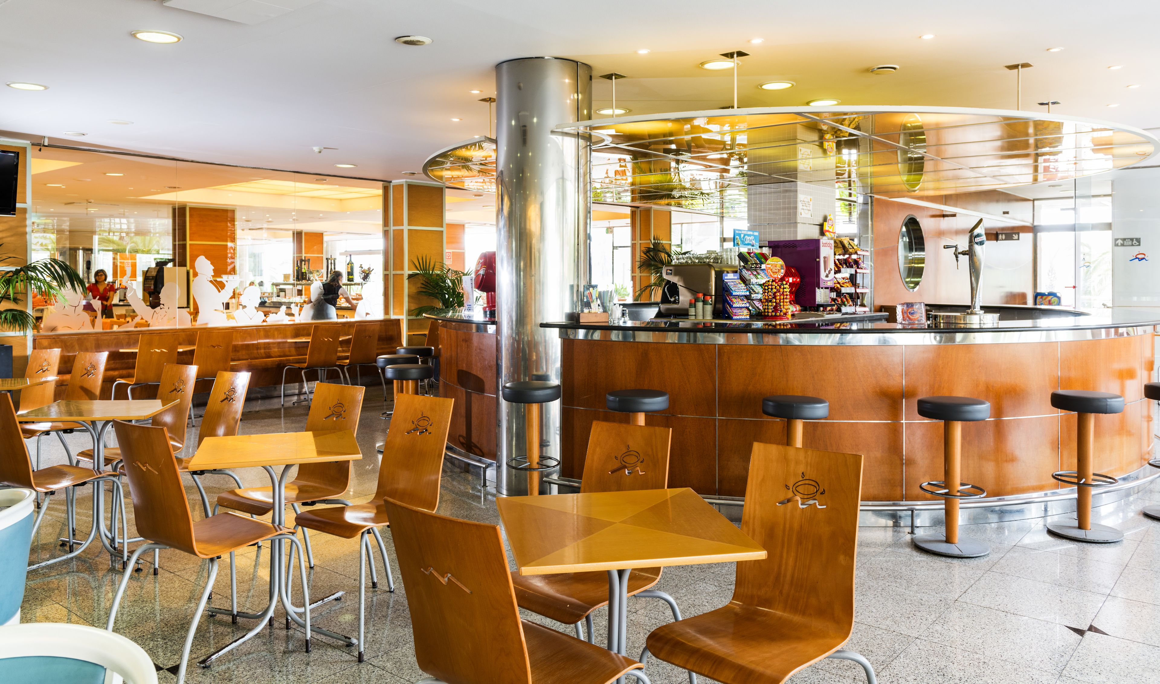 Un restaurante moderno con mesas y sillas de madera, una barra central y un panel de vidrio que separa el comedor de la barra.