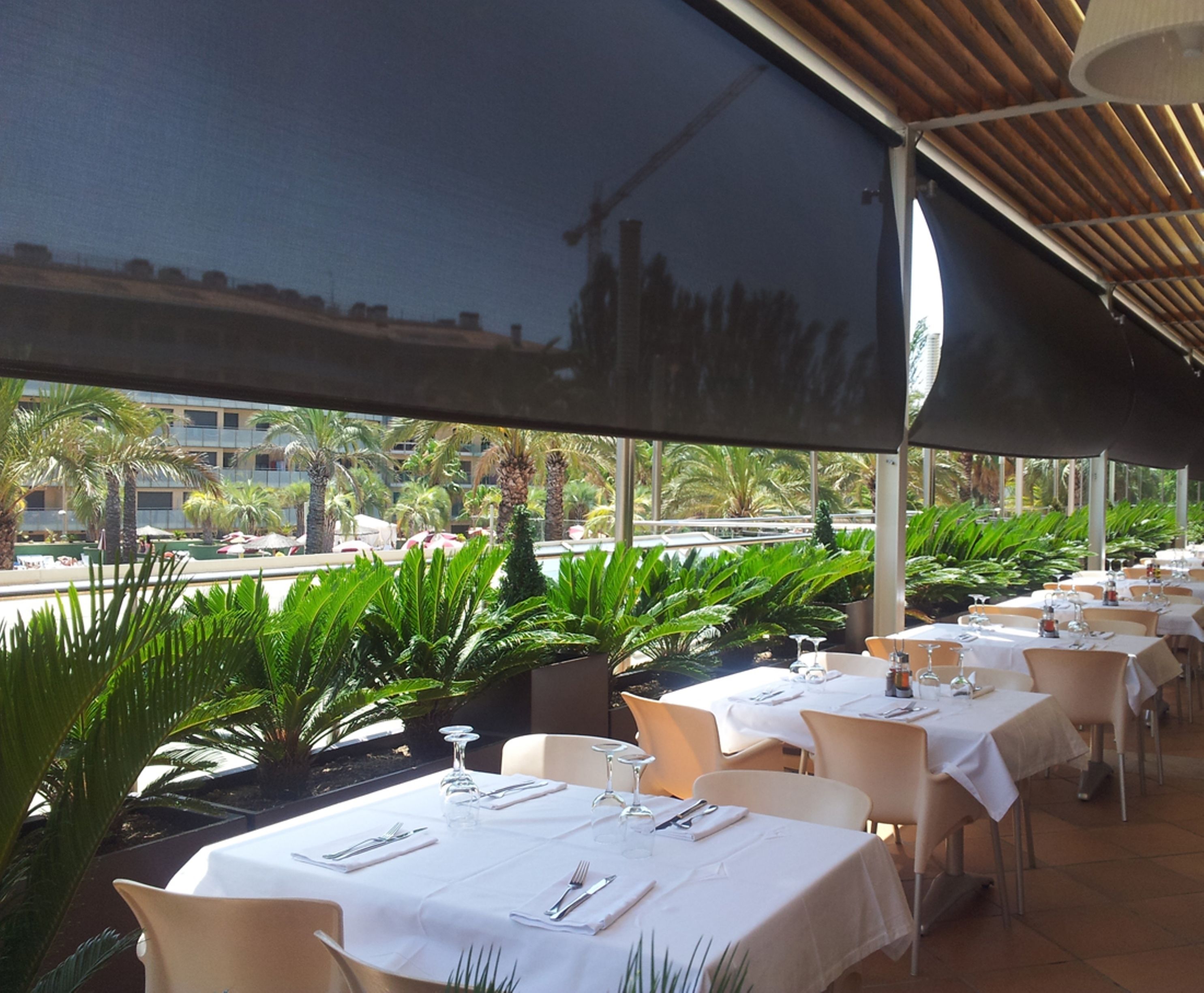 An outdoor dining area with tables set for a meal, surrounded by lush greenery and overlooking a pool and buildings.