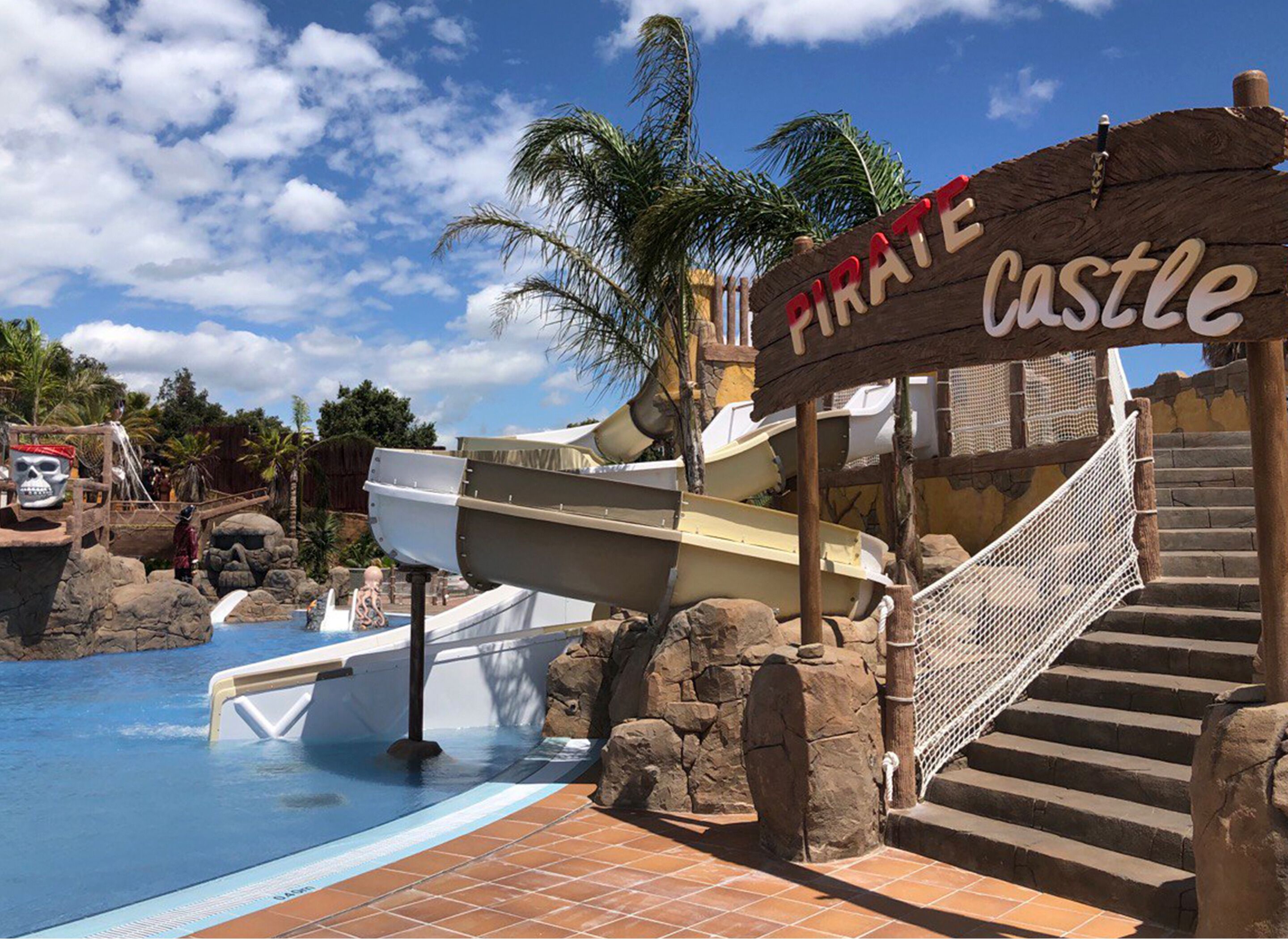 Ein wasserpark im Piratenthema mit einem Schild mit der Aufschrift 'Piratenburg', einem Pool, Wasserrutschen und einer Treppe, die zu einer Plattform führt.
