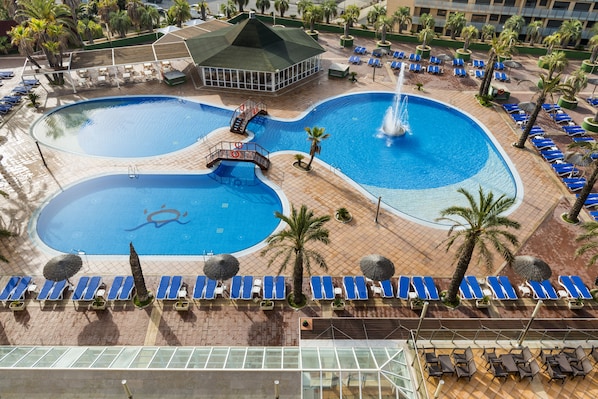 A resort with two large swimming pools, a covered area, and numerous sun loungers.