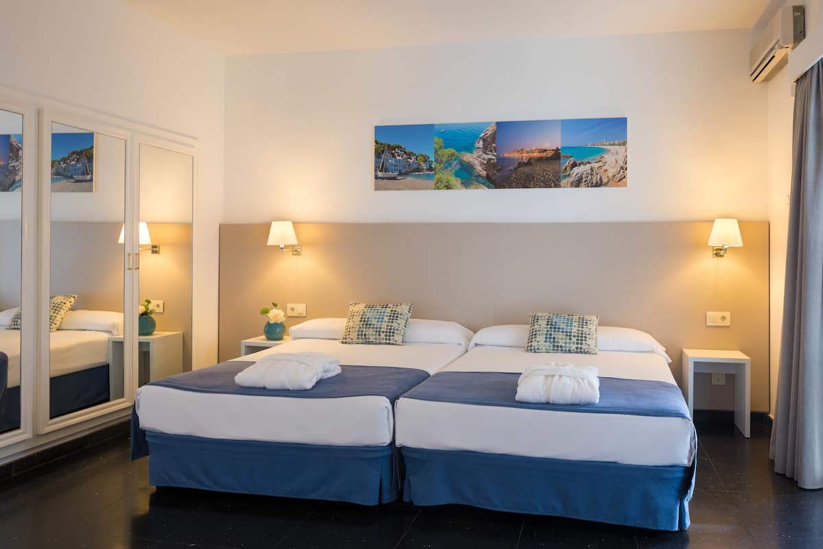 A double room with two beds, each with a blue bedspread, white pillows, and a white towel. There are two bedside tables with lamps, a large mirror, and a wall-mounted picture of a coastal scene.